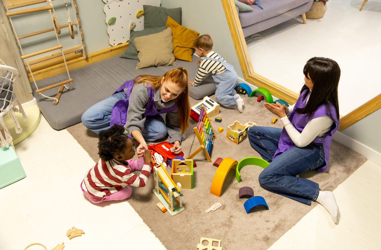 Two teachers play with toddlers on a soft rug in a modern preschool classroom with climbing equipment visible - indoor active-play zone in use