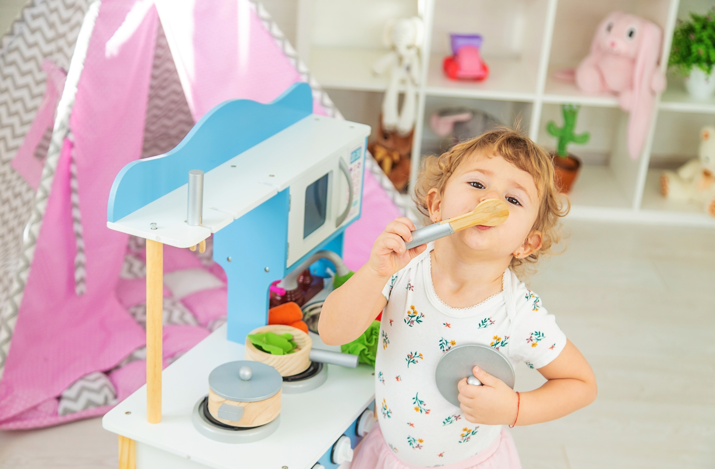 A toddler with curly hair plays with a wooden toy kitchen while holding a spoon to her mouth and a pot lid in her hand. Behind her, a pink play tent and shelves filled with various toys are visible in a bright, child-friendly room.