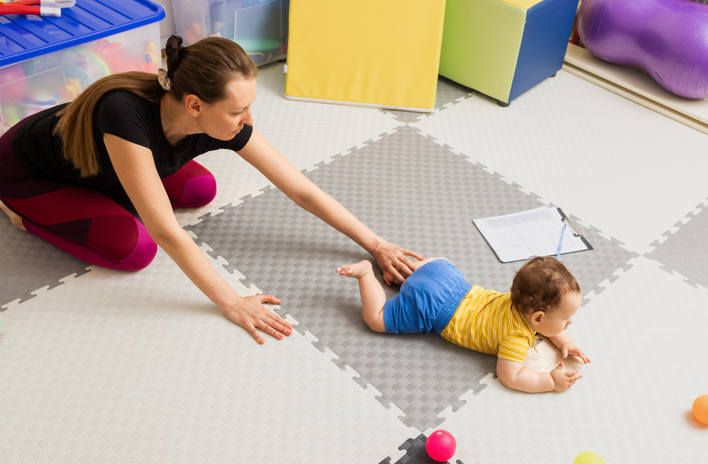 Adult kneeling on a foam play mat guiding a baby practicing crawling on a hard floor surface