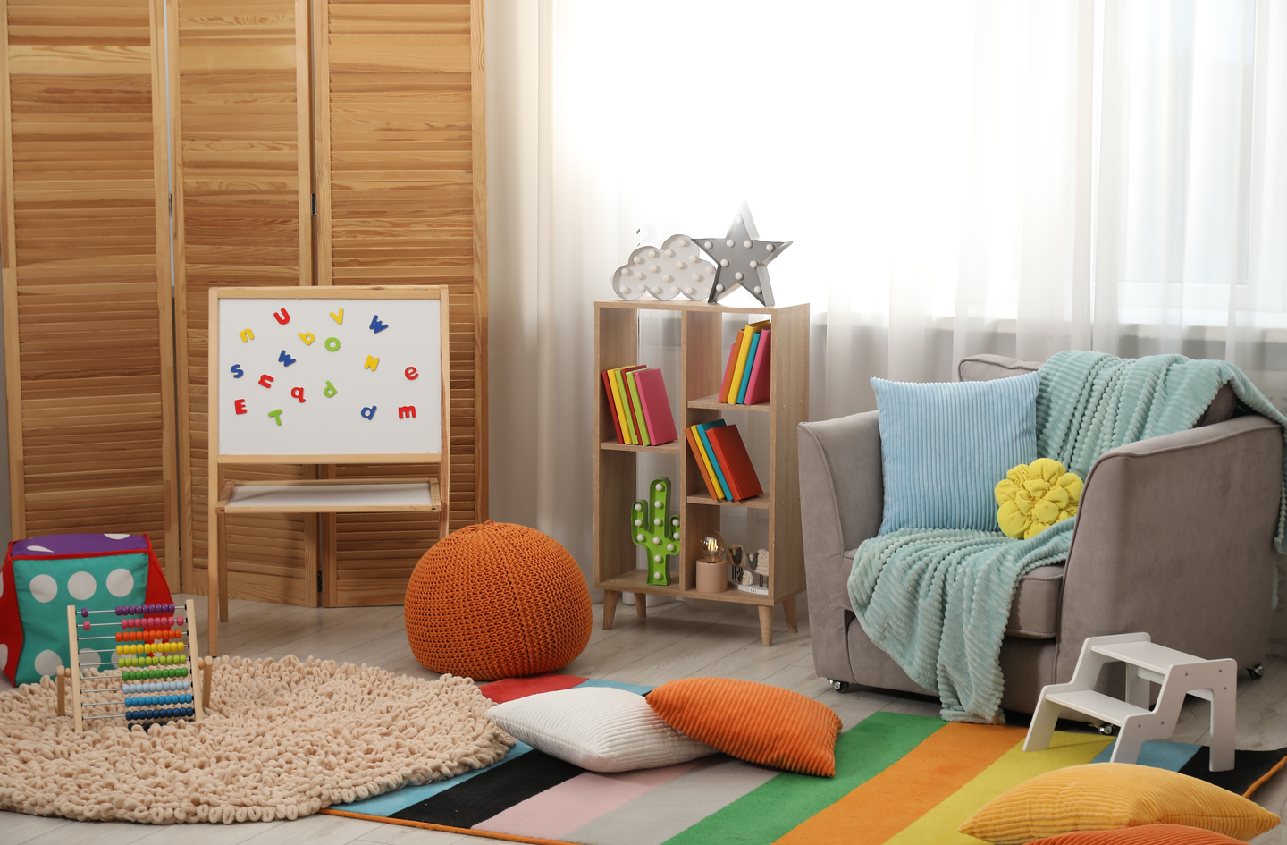 A bright playroom reading corner with a gray armchair angled toward the room, a striped rug marking the floor area and a wooden bookshelf filled with books.