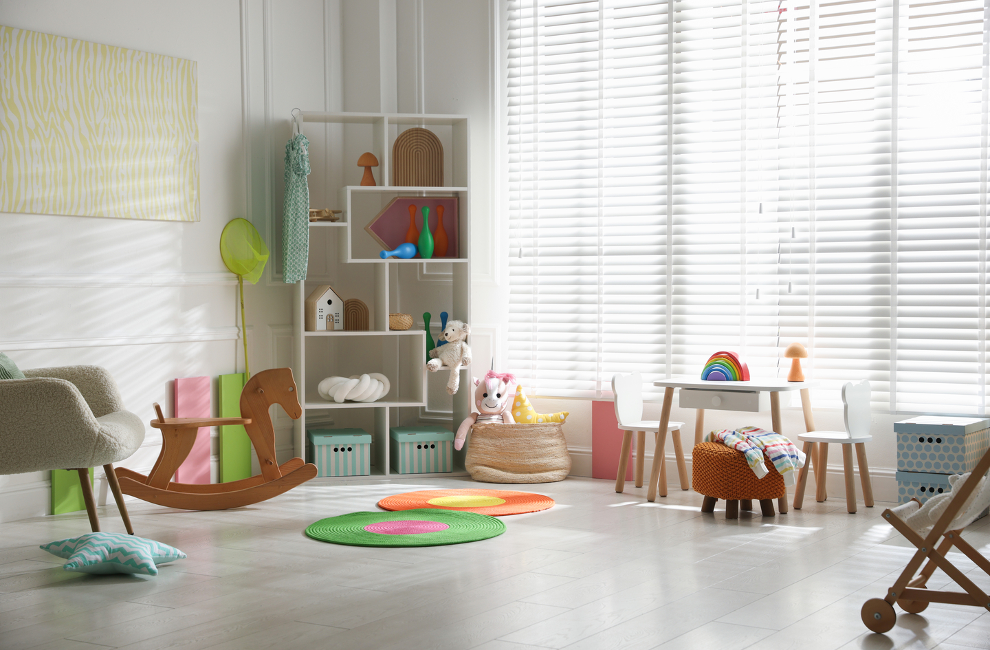A bright small playroom with a bookshelf and toys organized along the wall, a small table and chairs near the window, and a rocking horse on a colorful round rug in the open center area.