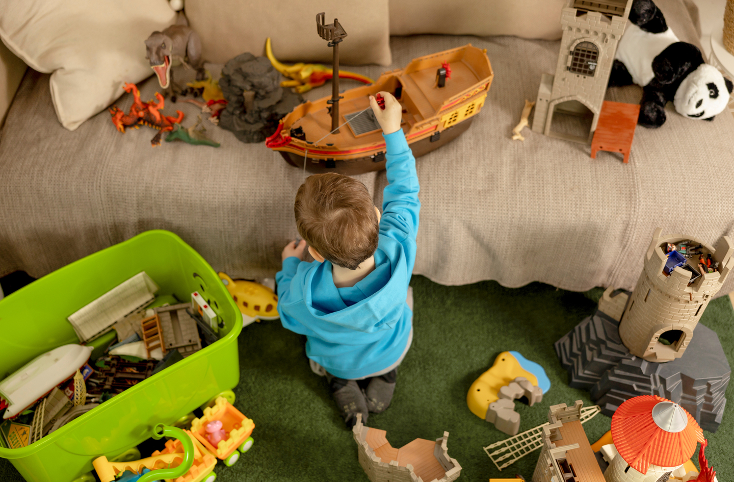 A young child in a blue hoodie plays on a green padded mat surface surrounded by toys including a pirate ship play set.