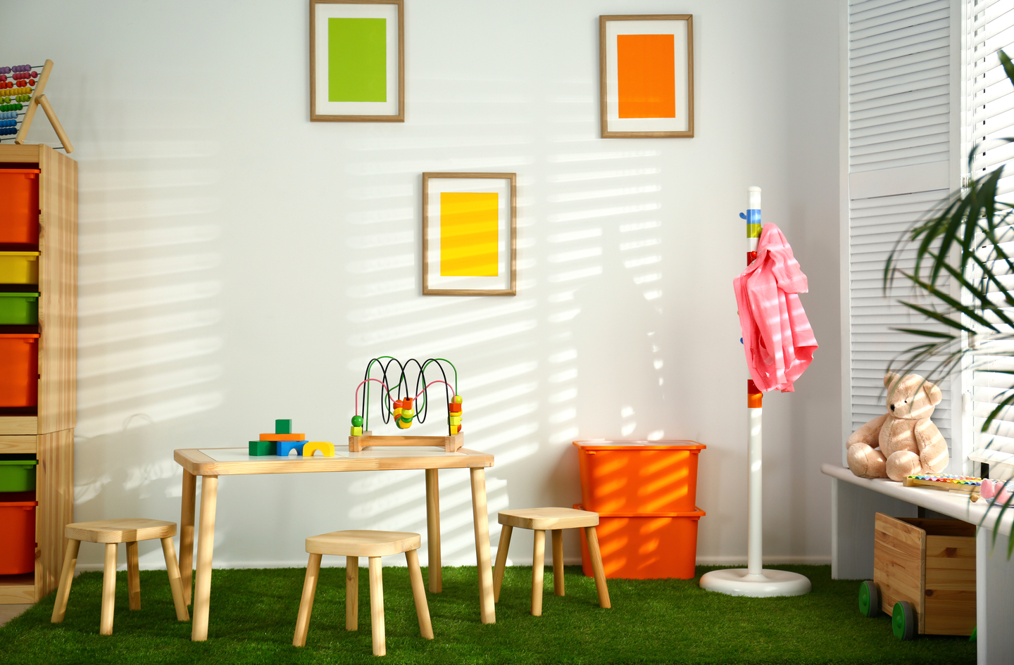 A brightly lit playroom with a thick green padded play mat covering the floor beneath a child-sized wooden table and stools.