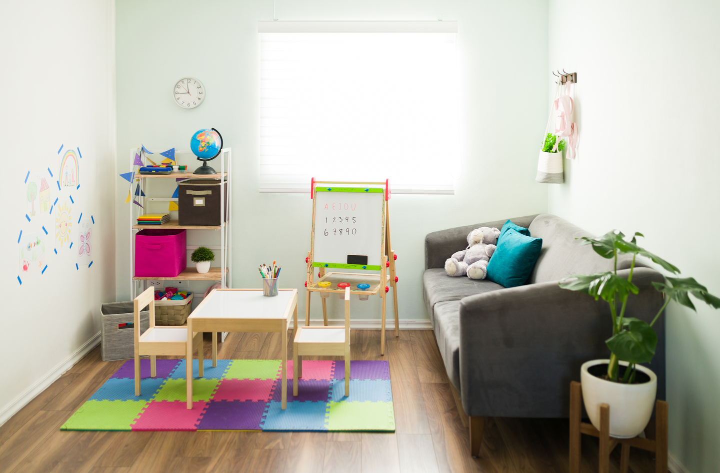 Small playroom showing distinct activity zones including a table and chairs on a foam mat, a tall storage shelf along one wall, and a couch for calm seating