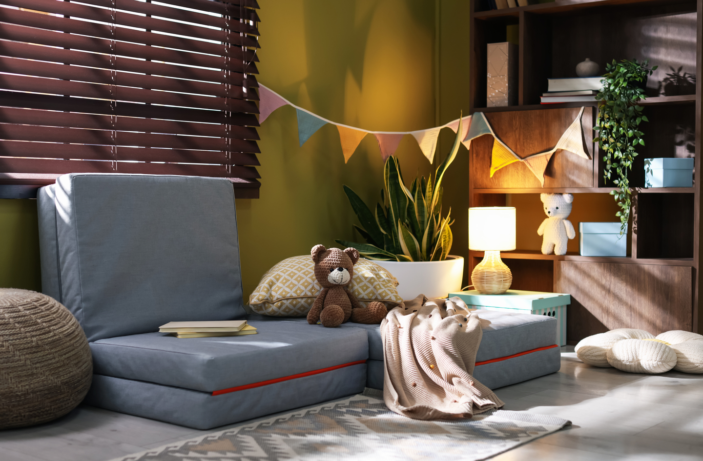 A grey floor sofa in a sunlit playroom corner with a wooden shelf holding books and a small lamp - a simple kids' reading nook setup