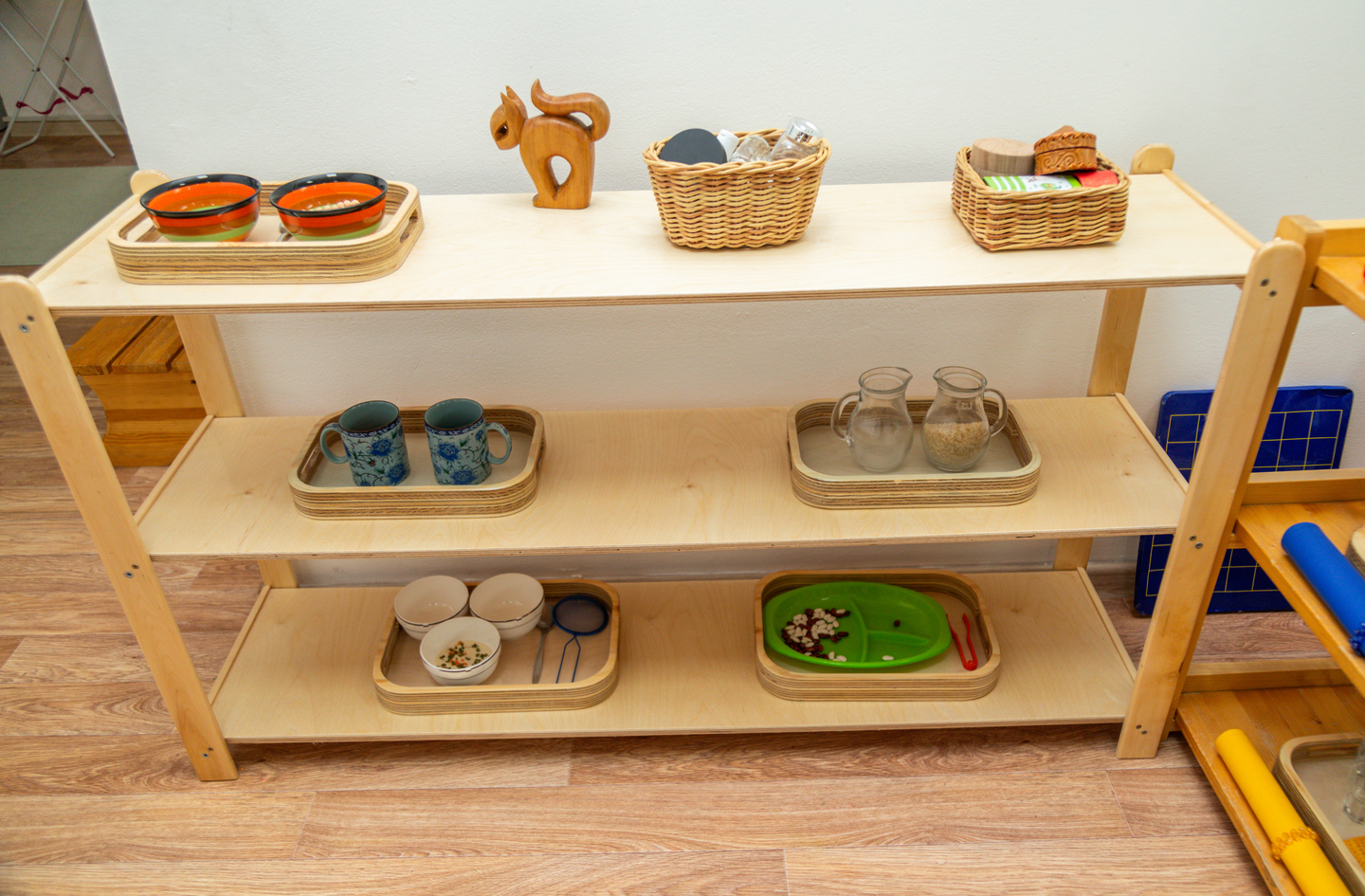 A wooden Montessori shelving unit displaying learning materials arranged in individual trays, including sorting activities, pitchers, and small containers
