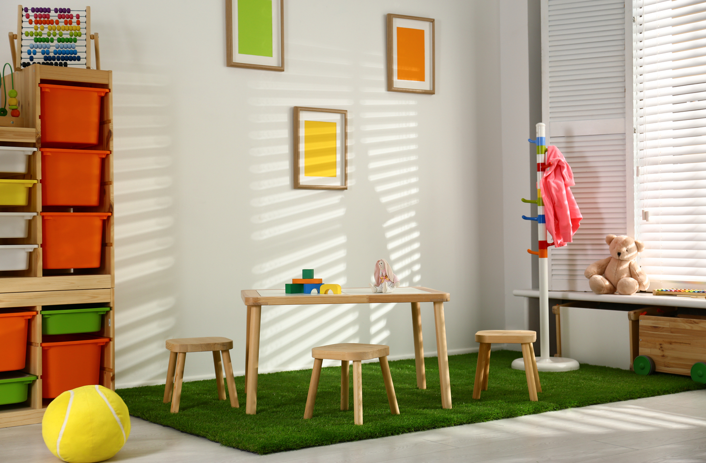 Small playroom ideas in action: a bright room with a central wooden table on a grass rug, colorful storage bins along the wall, a coat rack, and an open center floor that keeps movement paths clear.