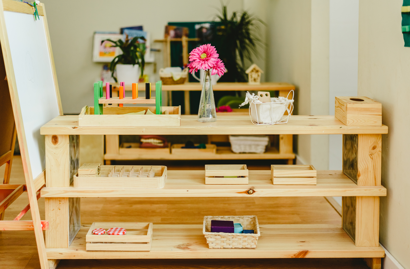 A low wooden shelving unit displays small trays, wooden pegs, a vase with flowers, and a standing whiteboard in a bright organized playroom