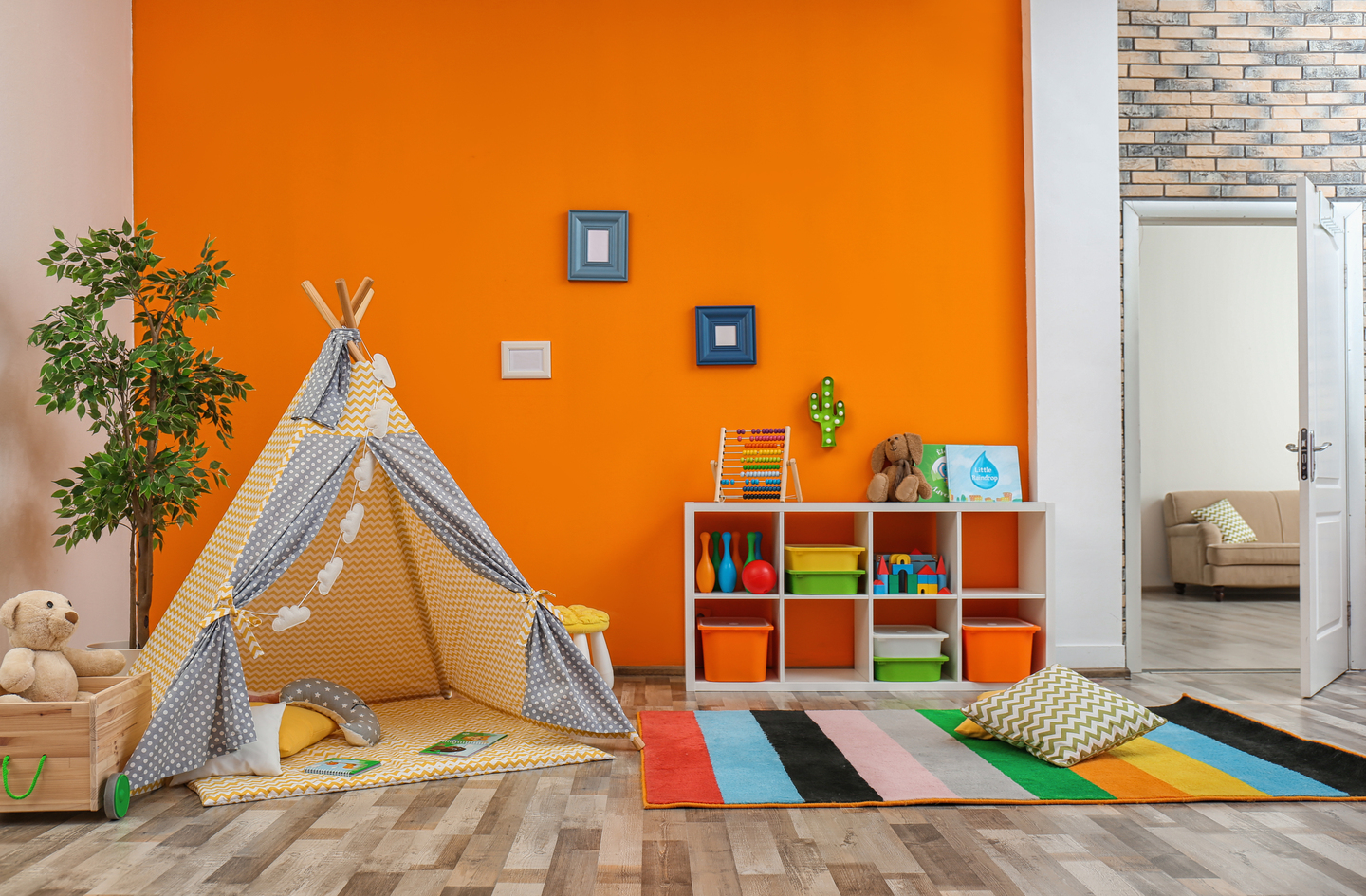 Small-space playroom layout with a teepee, a multi-colored rug, and a white toy shelf against an orange wall