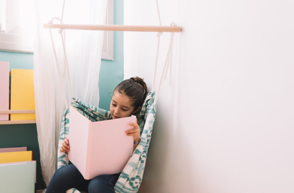 One of several corner reading nook ideas: a young girl reads a pink book in a patterned fabric hanging chair tucked into a playroom corner beside a wooden shelf of colorful books.
