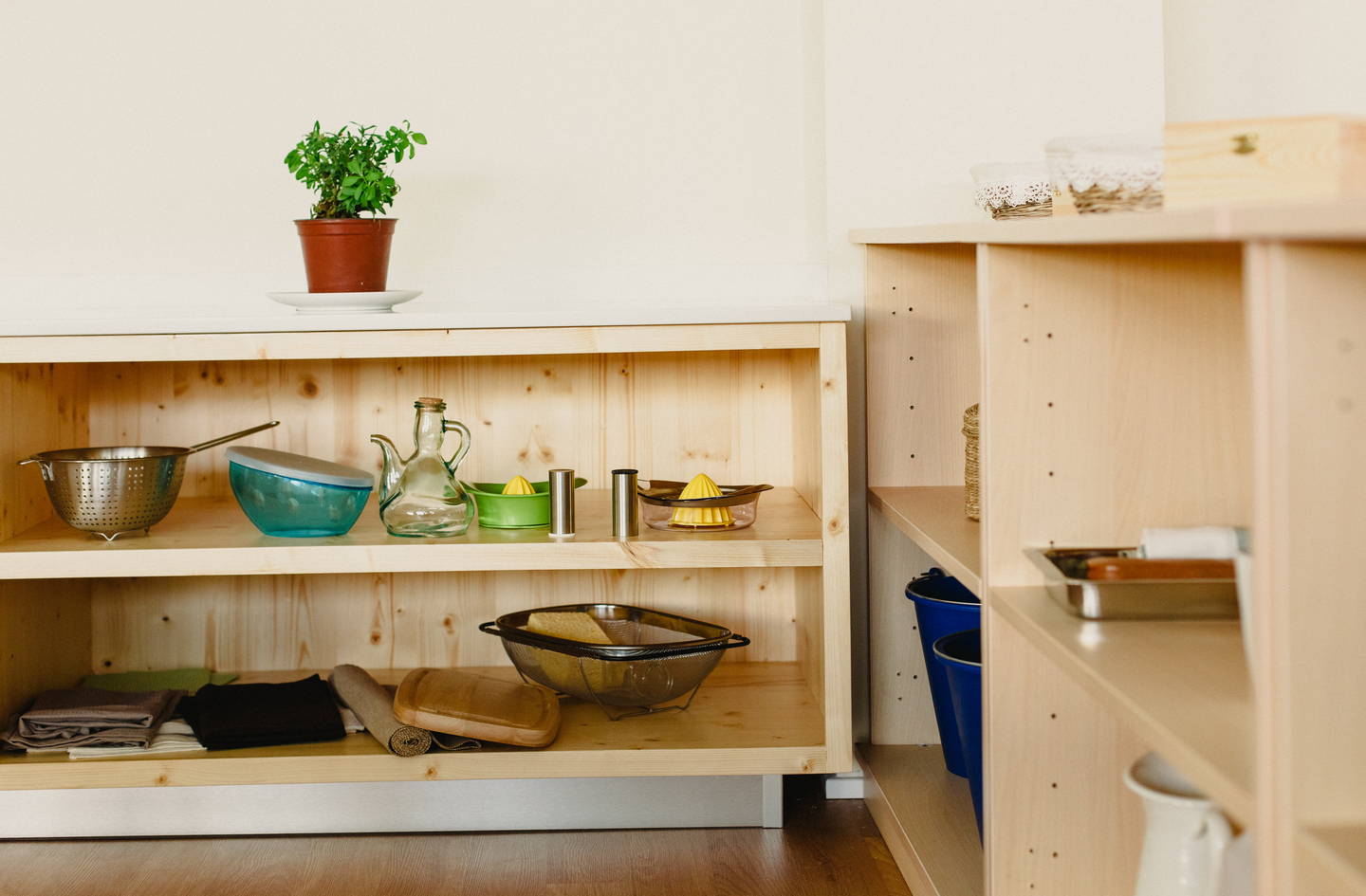 A low wooden shelf holds everyday kitchen tools, including bowls, a carafe, citrus juicers and folded cloths, with blue buckets and a metal tray on a second shelf beside it.