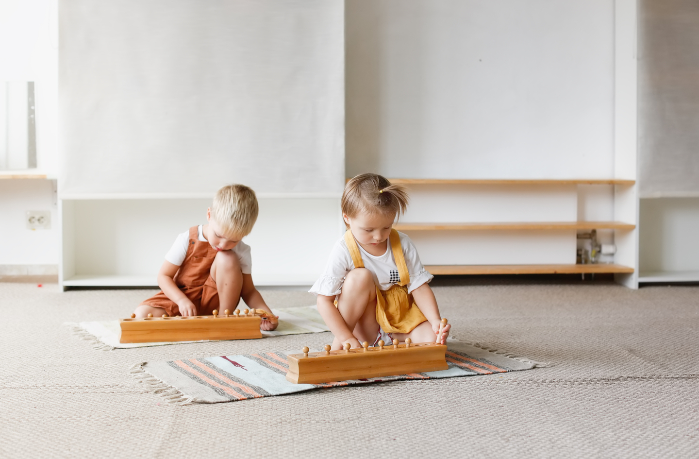 Two children working on individual floor rugs with Montessori cylinder block sets in a minimalist playroom