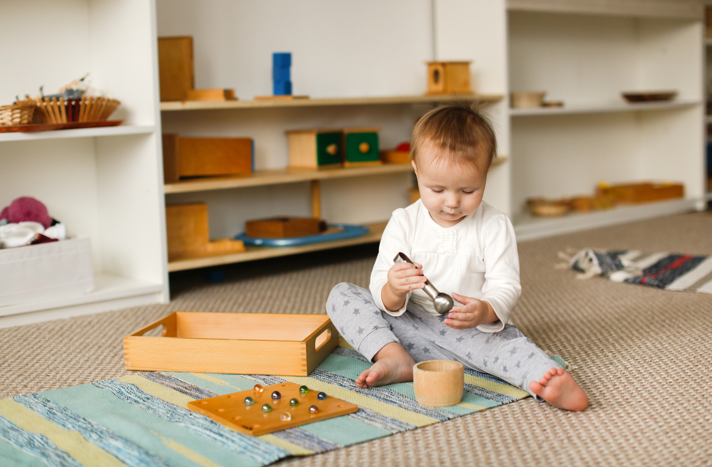 A toddler on a colorful rug uses a small metal scoop beside a wooden marble board, two simple sensory play toys for toddlers suited to one scooping activity.